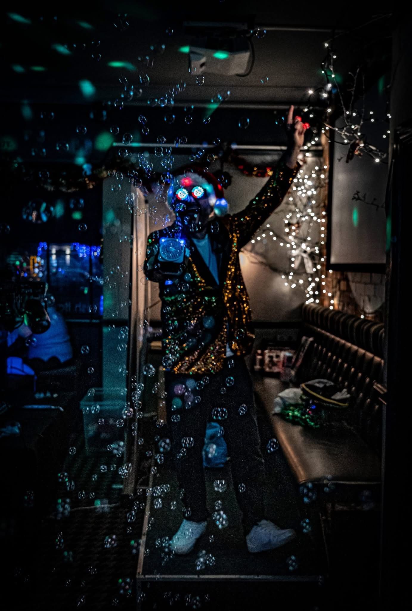 Sequin-jacketed host in light-up glasses surrounded by floating bubbles and fairy lights during a Christmas party night