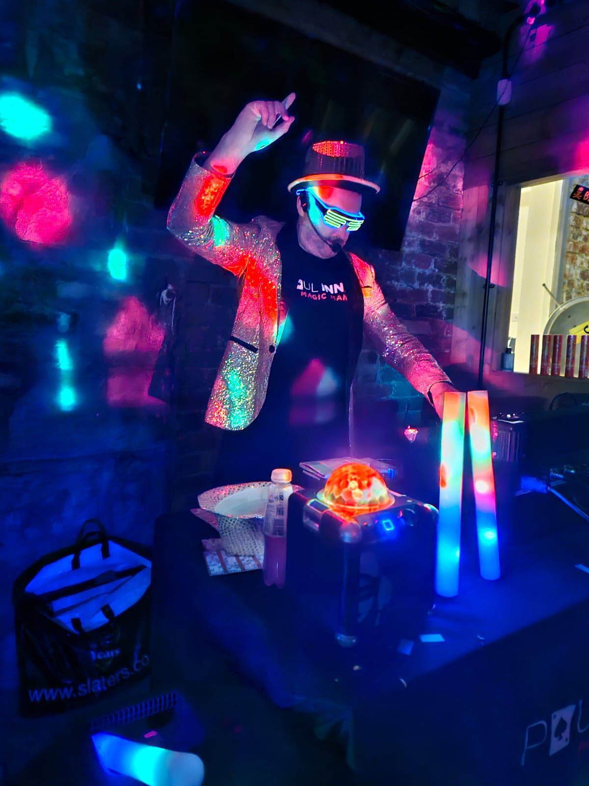 Host in a sequin jacket and light-up glasses dancing behind a disco ball speaker and glow sticks at Christmas Bonkers Bingo