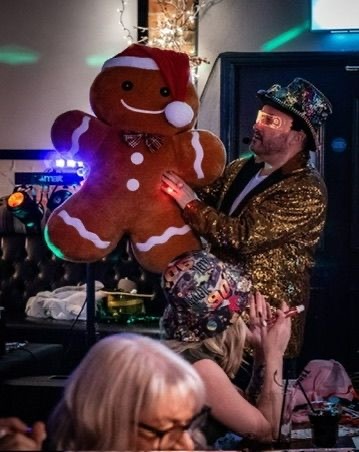 Host in a gold sequin jacket holding a giant gingerbread man in a Santa hat at a Christmas party night